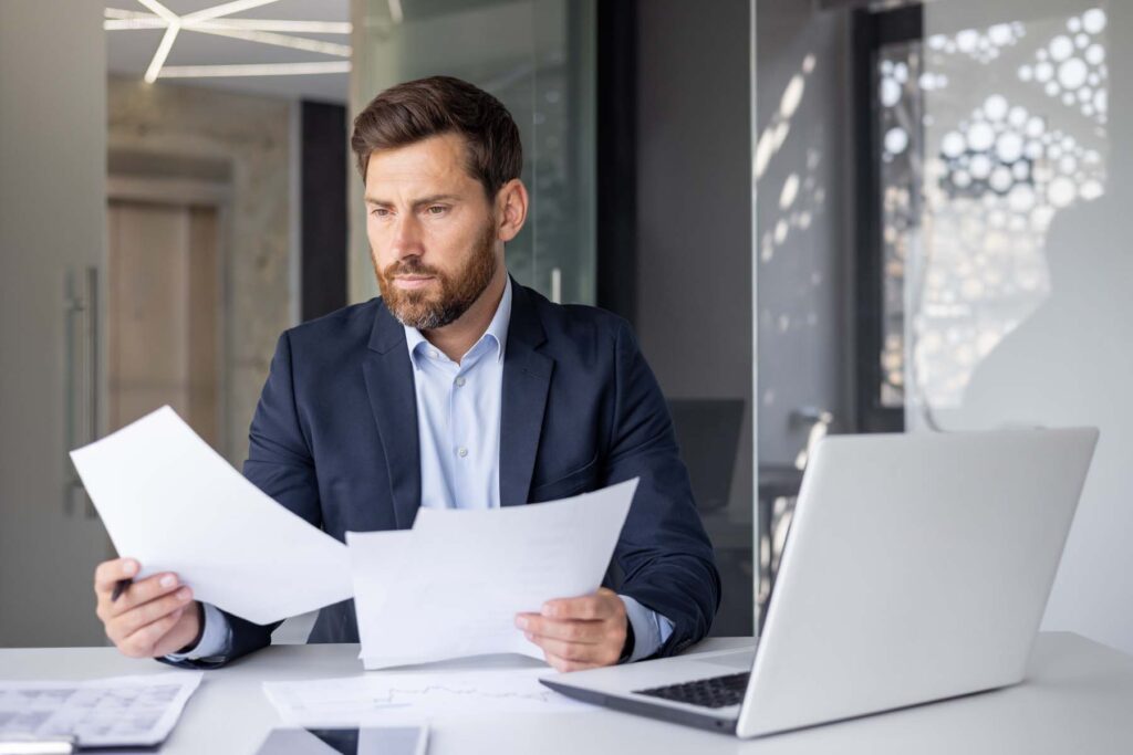serious and concentrated young man businessman sitting in the office at the desk with a laptop in a suit and working with documents and a project